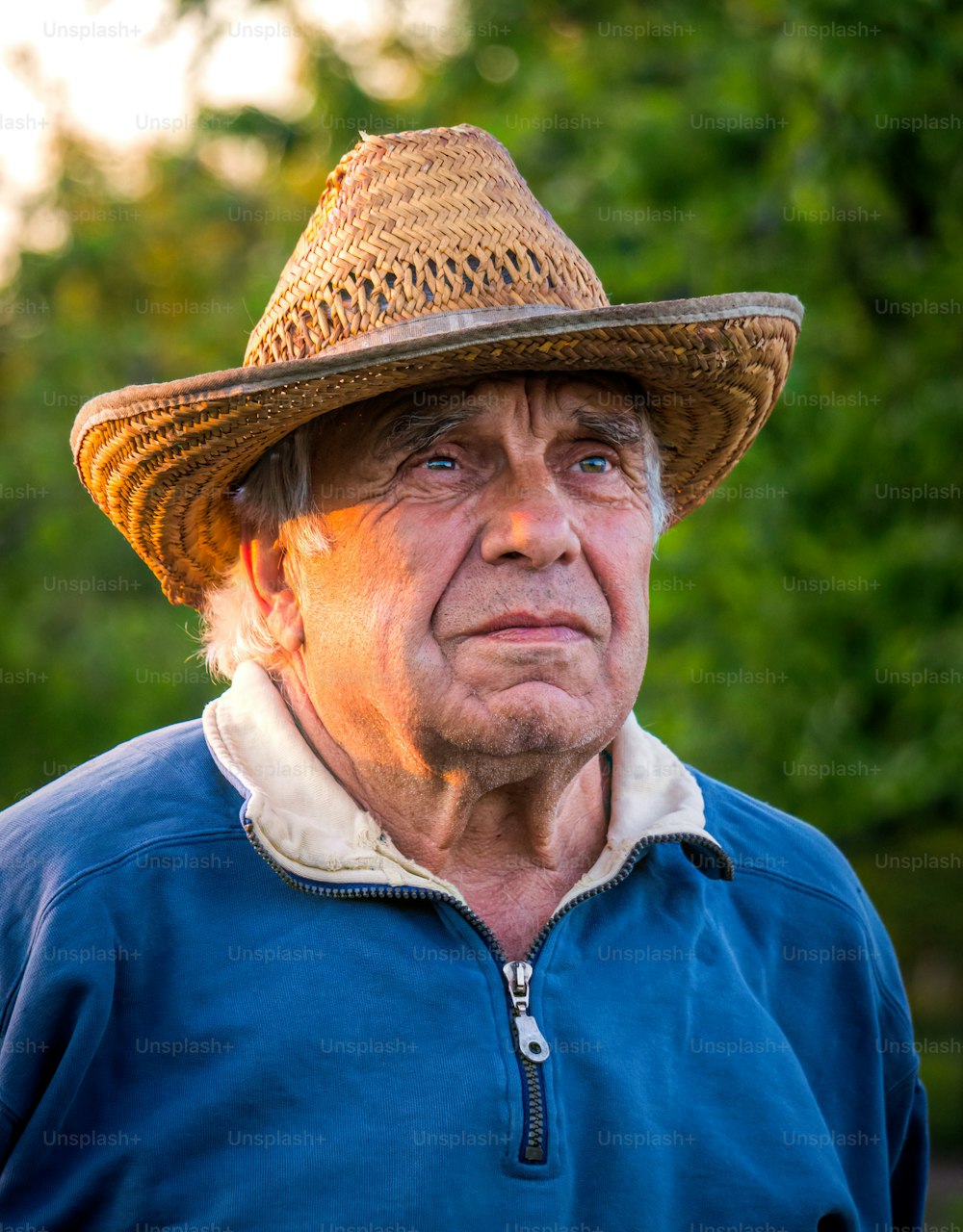 Ritratto di un anziano uomo dai capelli grigi in un cappello di paglia su uno sfondo di un soleggiato giardino primaverile e cielo al tramonto. Tranquilla vita di villaggio in pensione