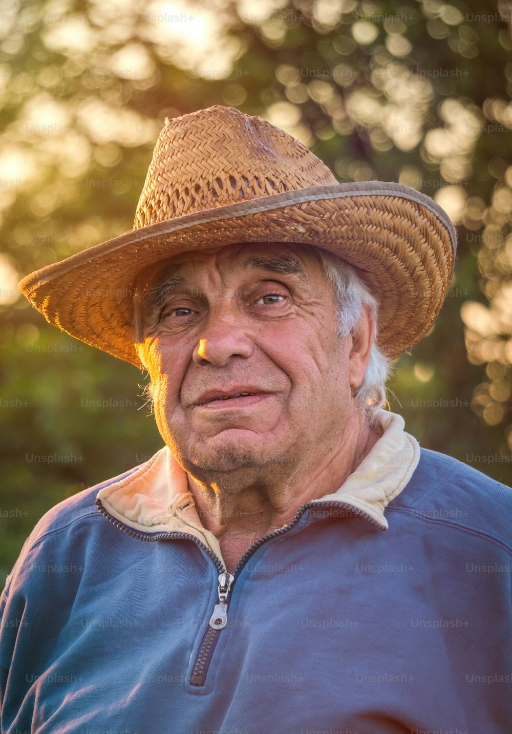 Portrait of an elderly gray-haired man in a straw hat on a background of a sunny spring garden and sunset sky. Peaceful retired village life