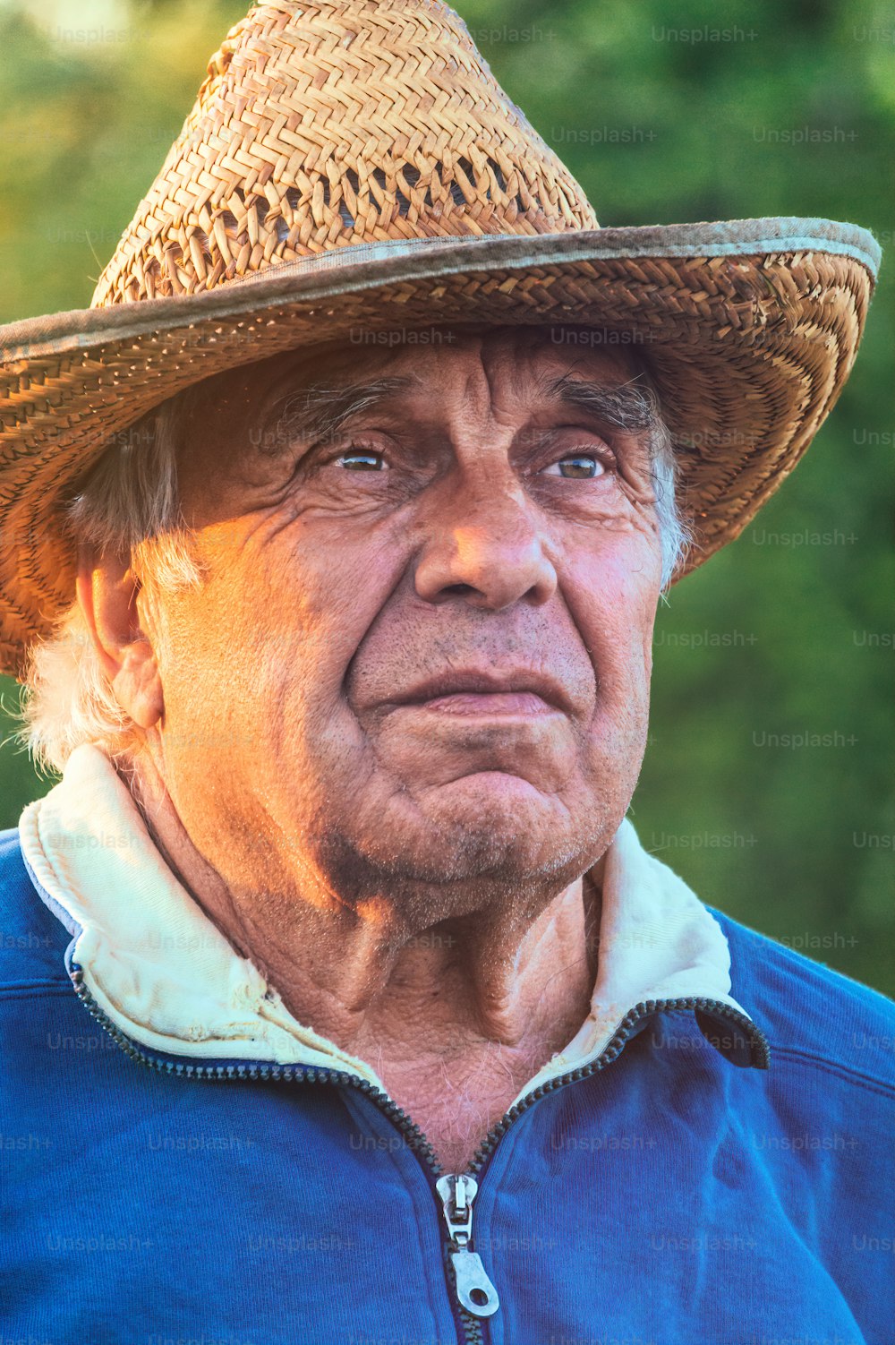 Retrato de um homem idoso de cabelos grisalhos em um chapéu de palha em um fundo de um jardim ensolarado da primavera e do céu do pôr do sol. Vida pacífica da aldeia aposentada