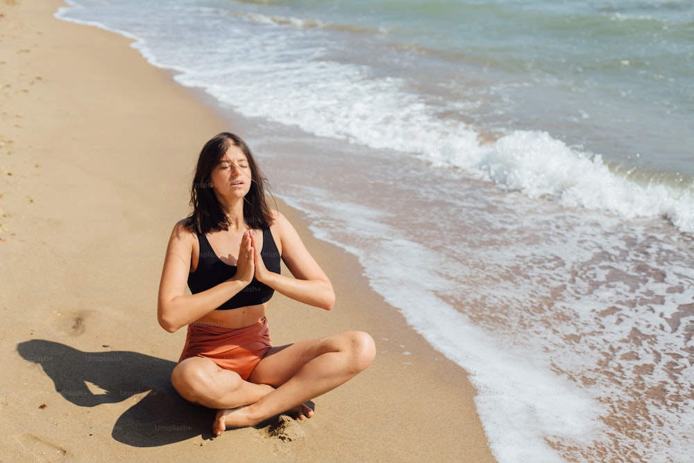Young beautiful woman practicing yoga on the beach, sitting on sand and meditaning. Mental health and self care concept. Happy girl relaxing on seashore on summer vacation