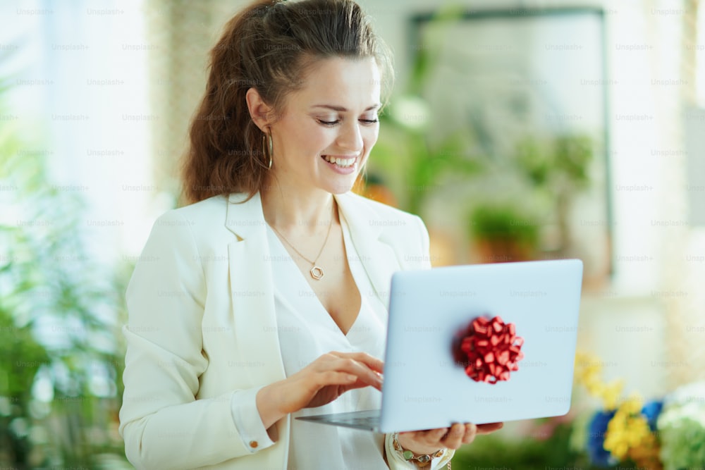 smiling trendy 40 years old housewife in white blouse and jacket using gift laptop with red bow in the modern living room in sunny day.