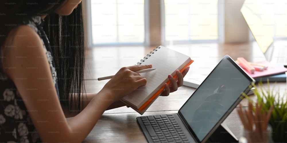 Cropped of young entrepreneur woman taking notes while sitting in front her computer tablet with keyboard case that putting on wooden working desk over modern sitting room windows as background.
