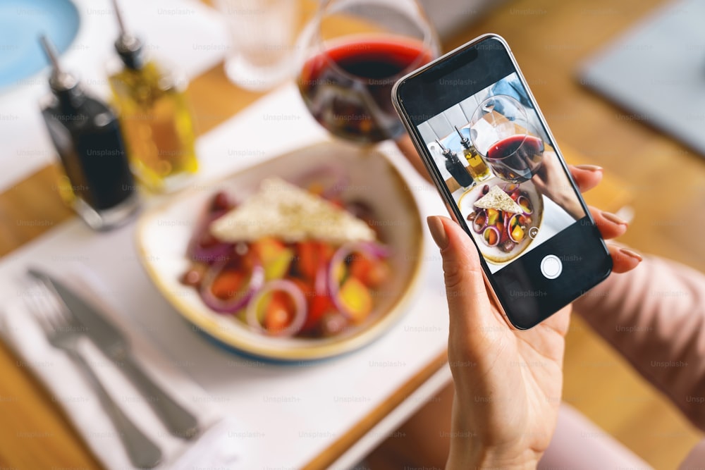 Gros plan de mains féminines tenant un smartphone et photographiant une salade fraîche et une boisson alcoolisée dans un café