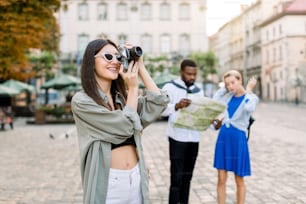 Pretty Caucasian brunette girl tourist in casual clothes taking photo in the city. African man and beautiful blond girl looking at the city map for some interesting places, standing on the background.