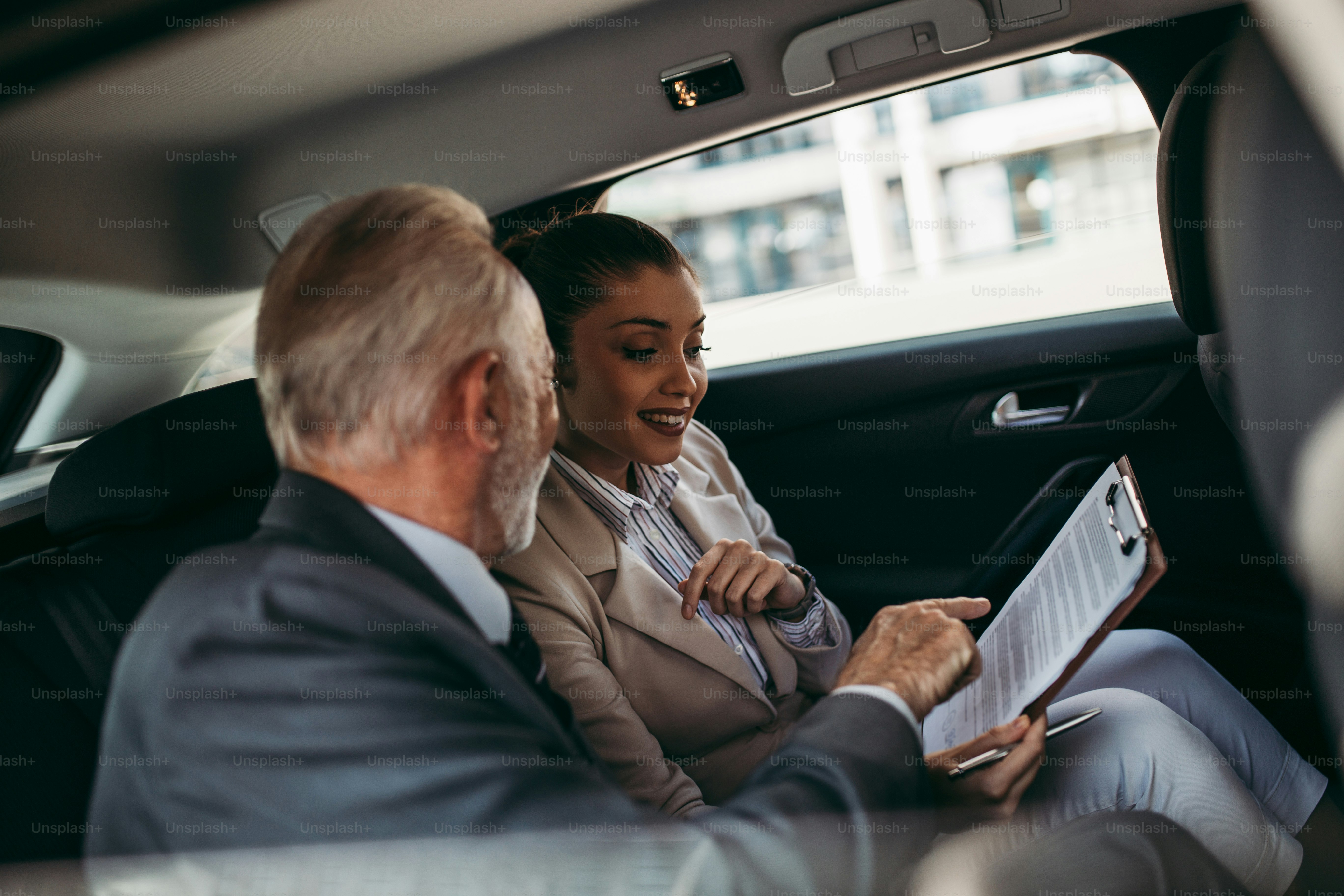 Bel homme d’affaires senior et sa jeune collègue ou collègue assise sur la banquette arrière d’une voiture de luxe. Ils parlent, sourient et utilisent un ordinateur portable et des téléphones intelligents. Transport dans le concept d’entreprise d’entreprise.
