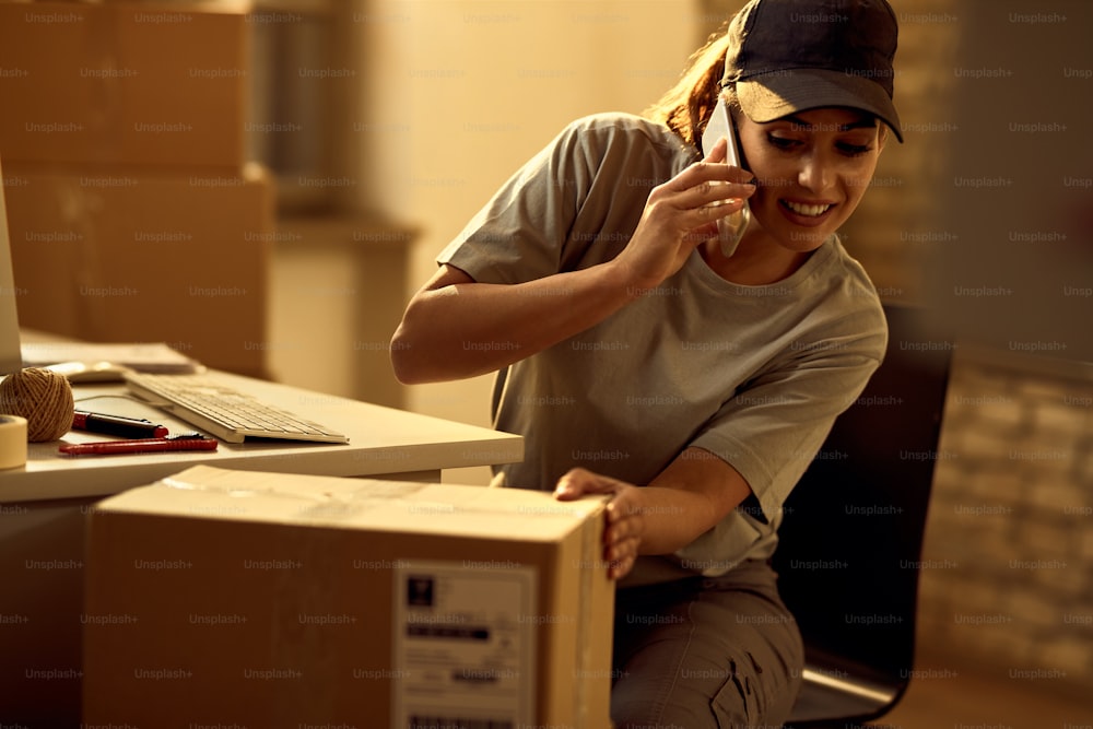 Happy female courier talking on the phone while checking packages in the office.