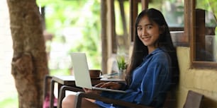 Mujer sonriente creativa que escribe en el teclado de la computadora portátil que se pone en su regazo mientras está sentada afuera en la silla de madera sobre el balcón de madera como fondo.