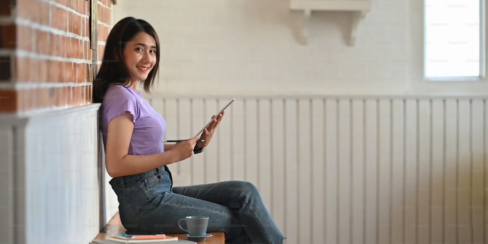 Photo of gorgeous young woman sitting with lean on brick wall while using a computer tablet and smiling over modern and vintage lounge as background.