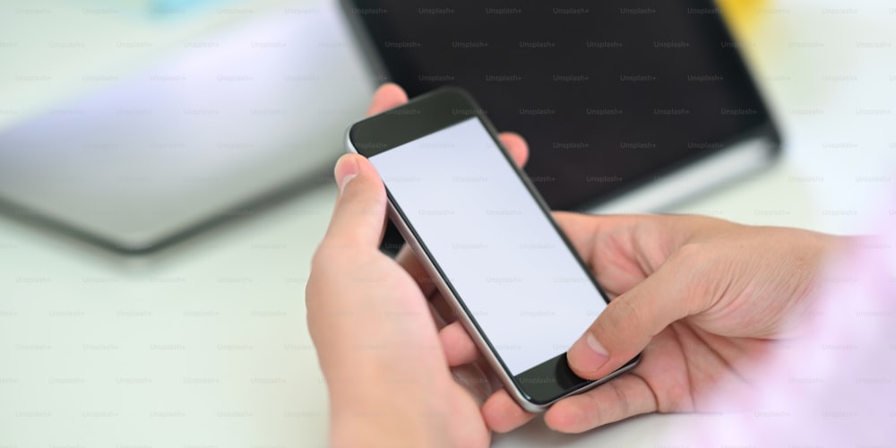 Cropped image of smart man's hands holding a white blank screen smartphone while sitting in front of computer tablet that putting on white working desk that surrounded by office equipment.