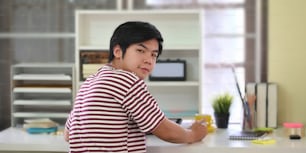 Photo of smart man in striped t-shirt working with computer tablet while sitting and turning back at the white working desk over comfortable working room as background.