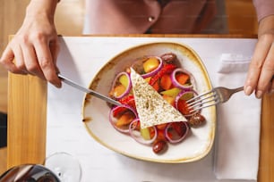 野菜、タマネギ、チーズの新鮮なサラダでテーブルに座っている間、フォークとナイフを持っている女性の接写