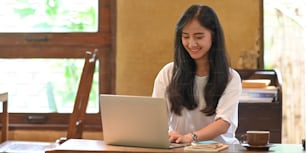Photo of attractive asian woman working with computer laptop that putting on wooden working desk that surrounded by coffee cup and stack of notebook over vintage living room as background.