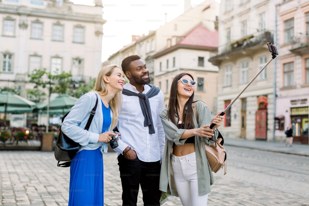 Multiethnic friends, tourists, young African man and two Caucasian women, wearing casual wear is exploring new city together, smiling and making photo on smartphone.