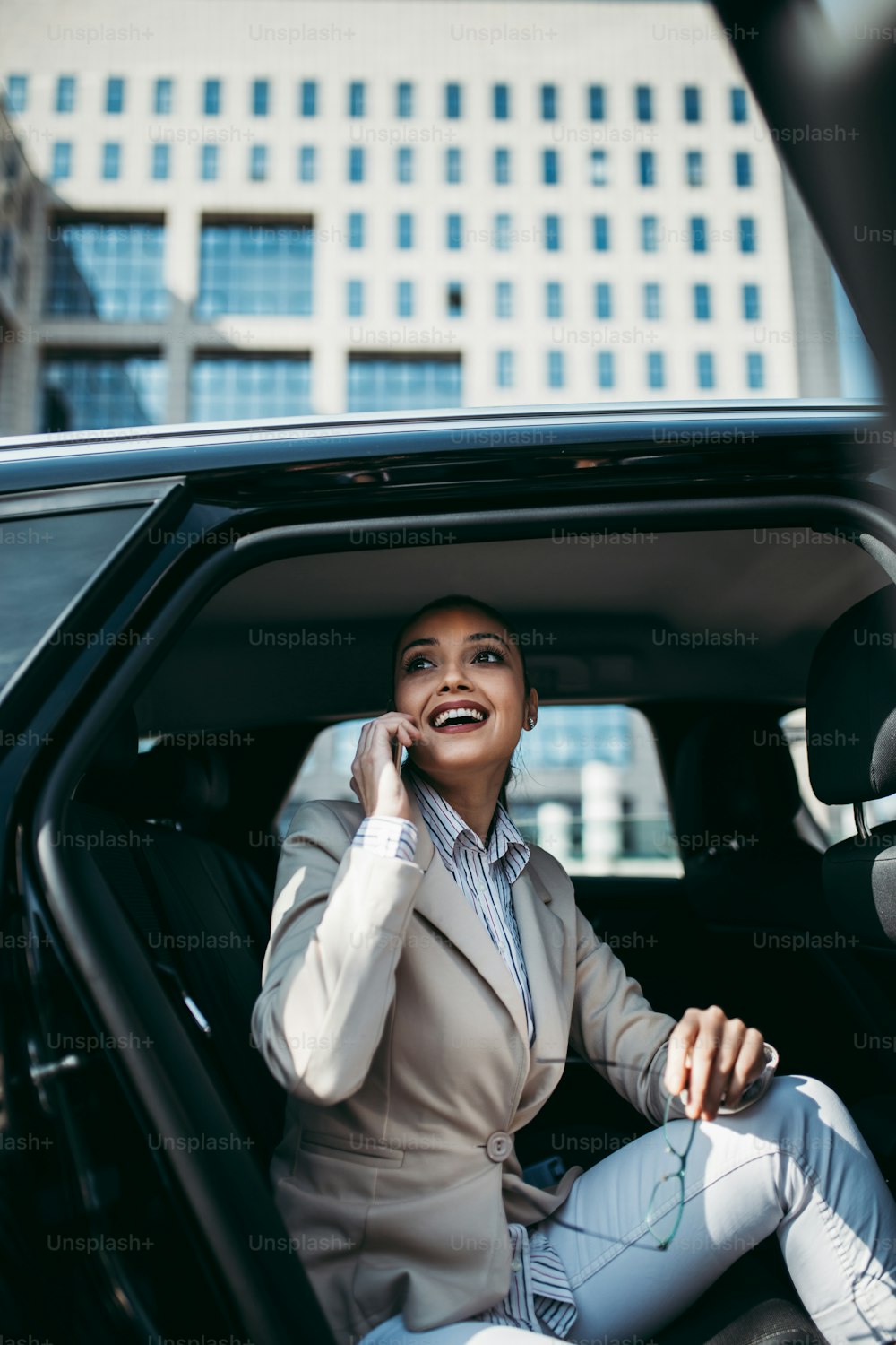 高級車の後部座席に座っている見栄えの良い若いビジネスウーマン。彼女はスマートフォンを使い、笑顔で外を眺めている。企業事業のコンセプトにおける輸送。