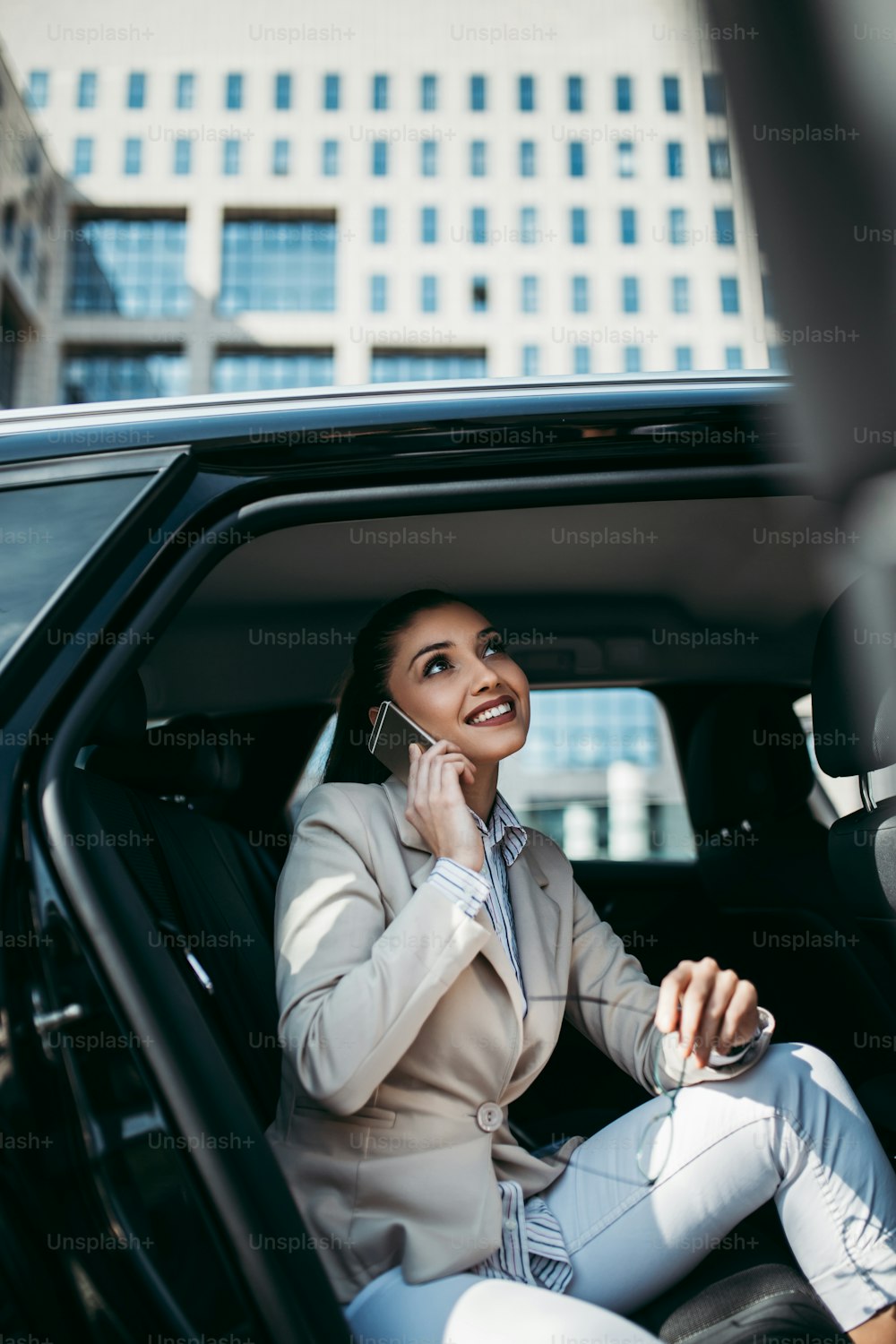 Joven y atractiva mujer de negocios sentada en el asiento trasero de un automóvil de lujo. Ella usando su teléfono inteligente, sonriendo y mirando hacia afuera. El transporte en el concepto de negocio corporativo.