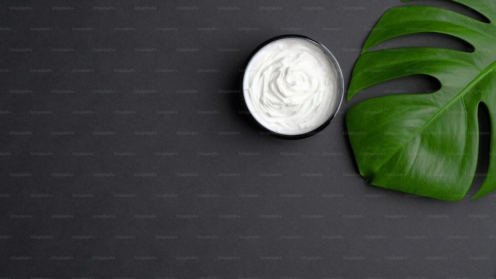 Jar with cosmetic cream and tropical monstera leaf on black background. Natural organic beauty product for body, hand, face skin treatment. Flat lay, top view. Skincare concept.