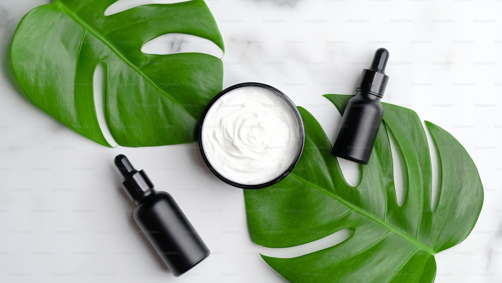 Set of natural cosmetic products on marble background top view. Flat lay black luxury dropper bottles with essential oil for body treatment, moisturizer cream and monstera leaves. Skincare concept