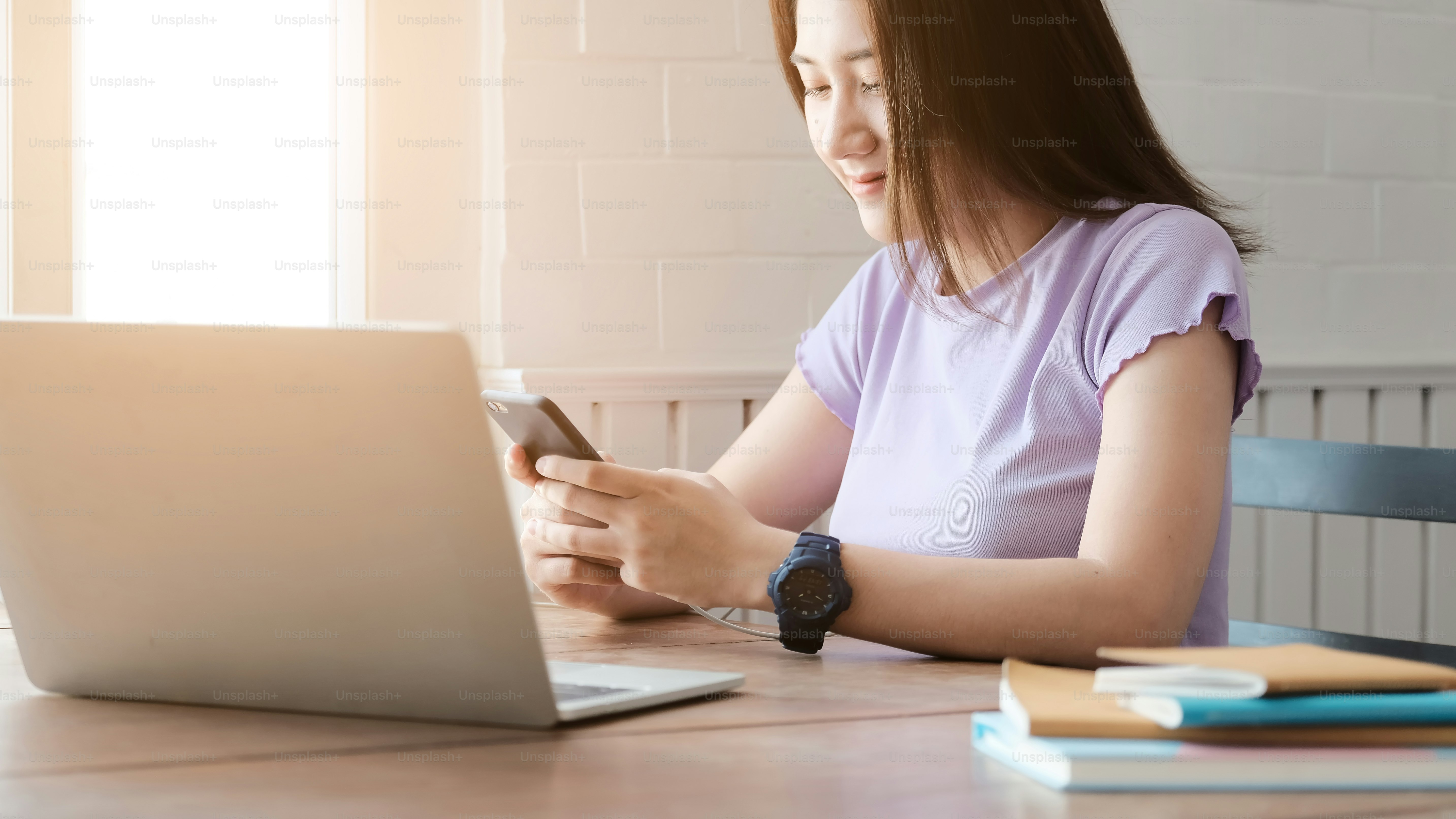 Gadis Asia yang bekerja di laptop dari rumah atau siswa yang belajar dari rumah atau pekerja lepas. Dia menggunakan smartphone untuk panggilan VDO.
