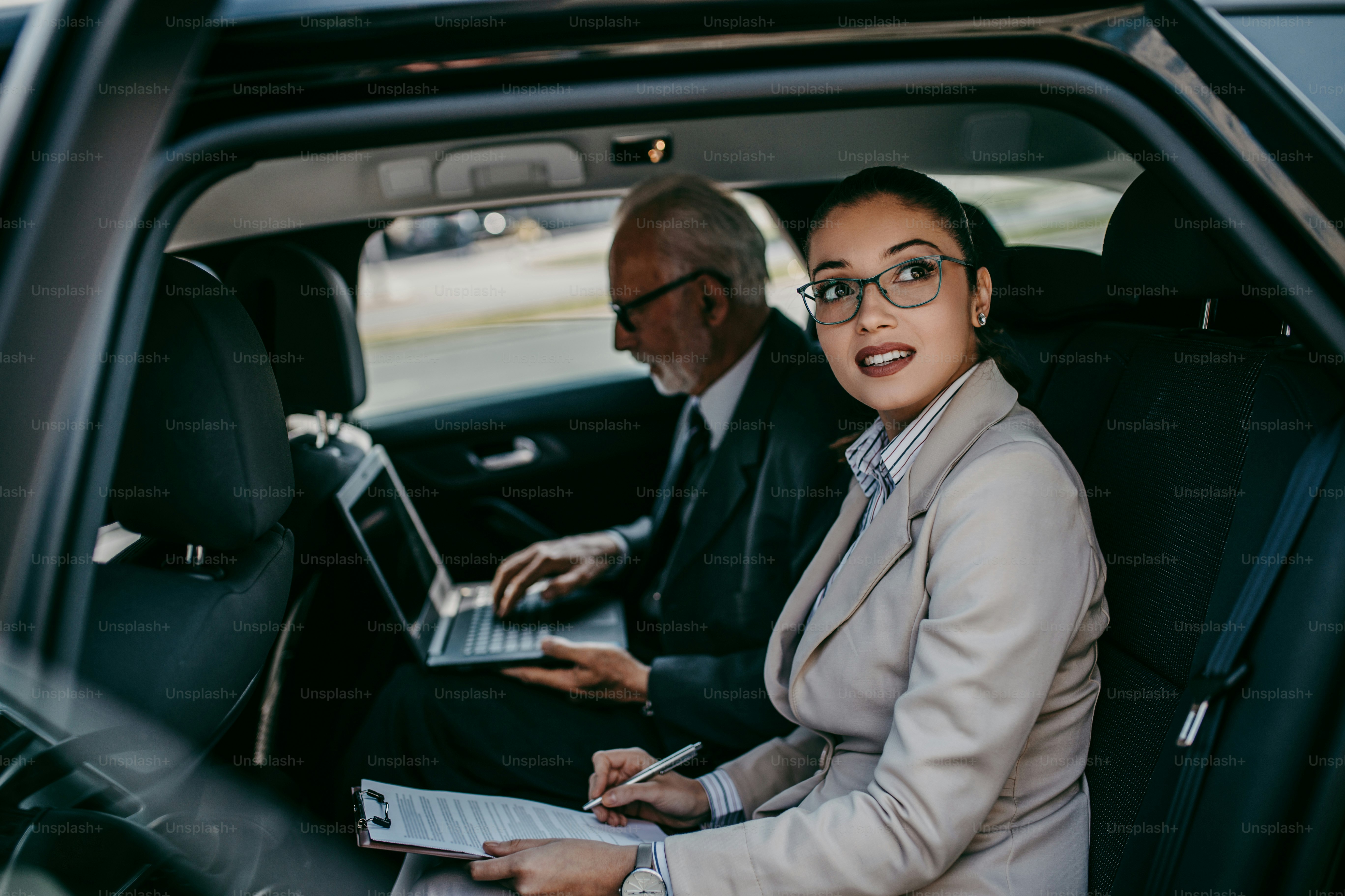 Good looking senior business man and his young woman colleague or coworker sitting on backseat in luxury car. They talking, smiling and using laptop and smart phones. Transportation in corporate business concept.