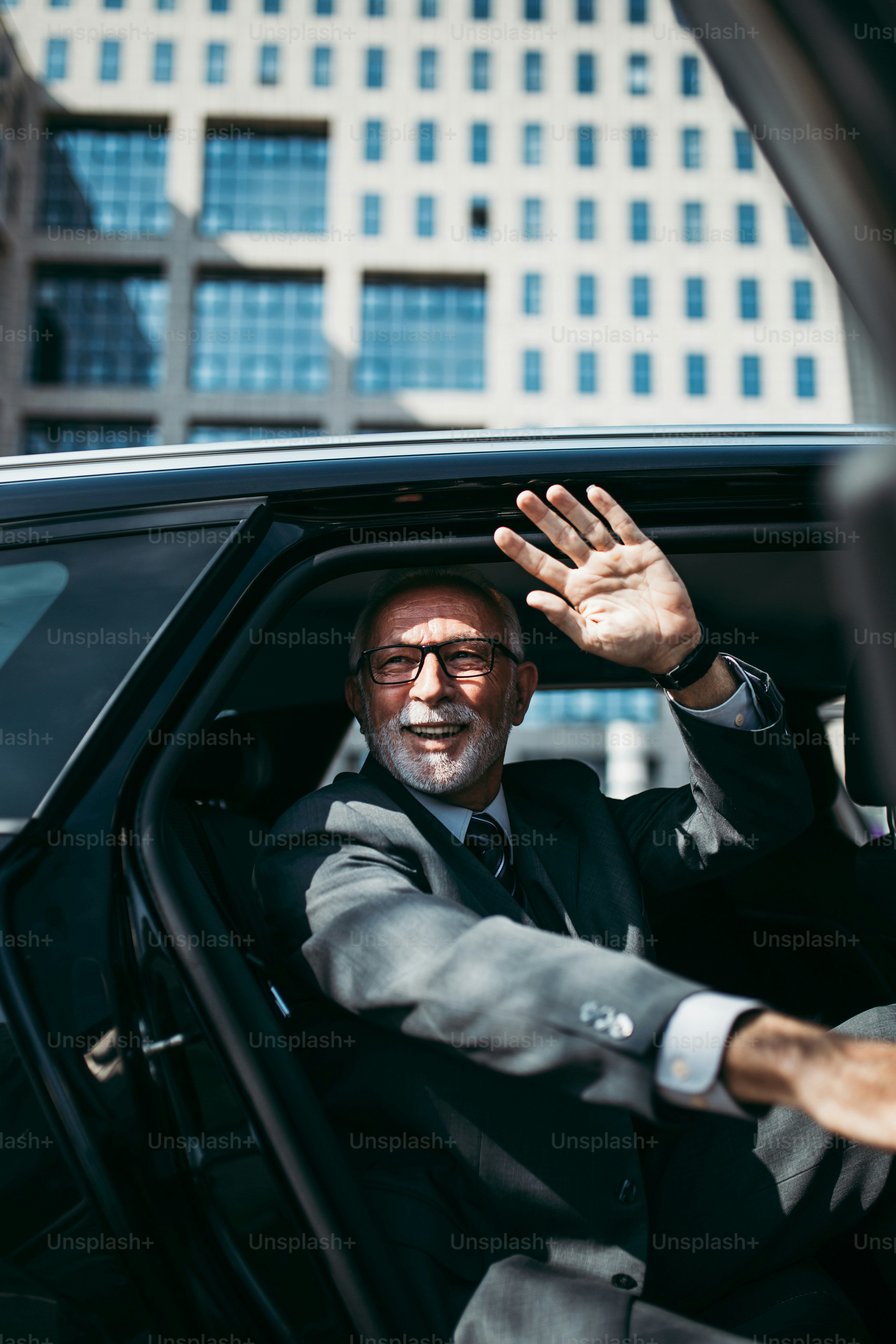 Good looking senior business man sitting on backseat in luxury car. He opens car doors and going or stepping out. Big busness bulding in background. Transportation in corporate business concept.