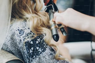 Hair curler. Hair stylist curling blonde hair with professional equipment closeup, morning preparations for wedding day. Bride in hair salon styling her hair