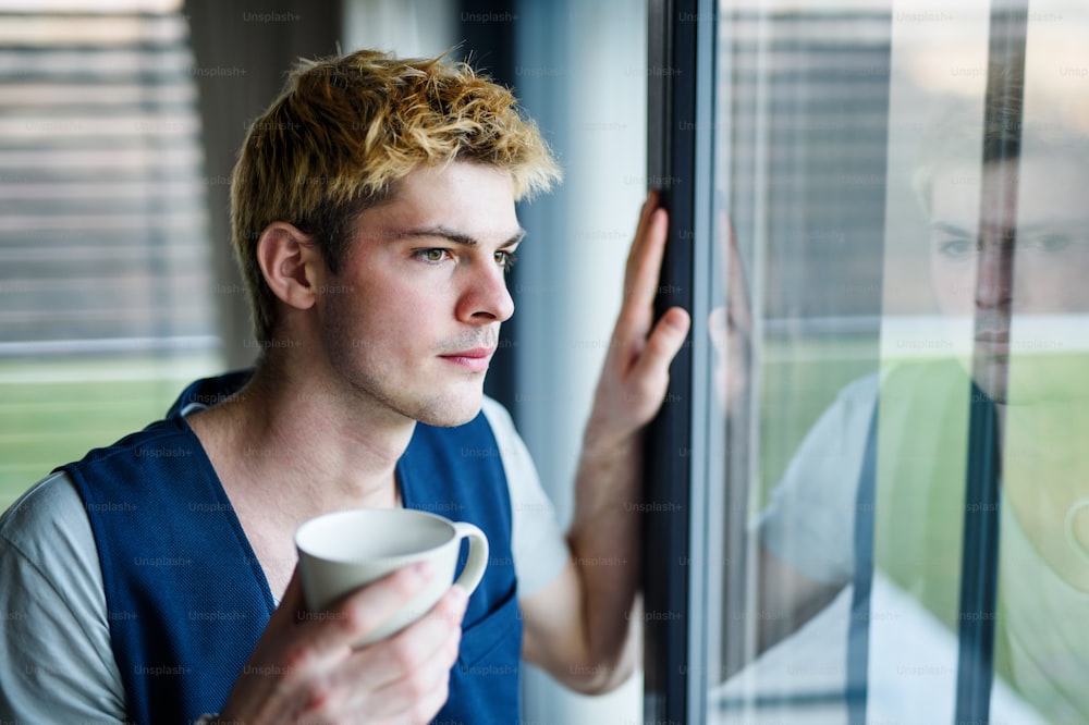 Young man with cup of coffee indoors at home, standing by window and looking out.