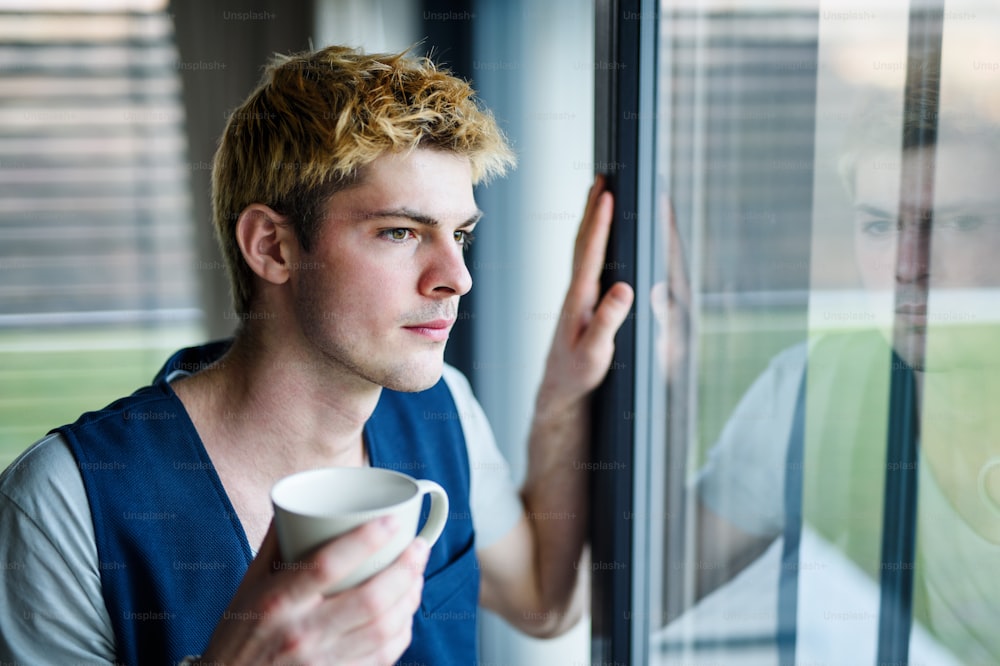 Junger Mann mit einer Tasse Kaffee drinnen zu Hause, am Fenster stehen und hinausschauen.