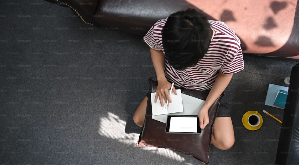 Immagine vista dall'alto dell'uomo creativo che lavora con la tavoletta del computer con schermo bianco vuoto che mette sulle ginocchia mentre prende appunti e si siede sul pavimento del salotto.
