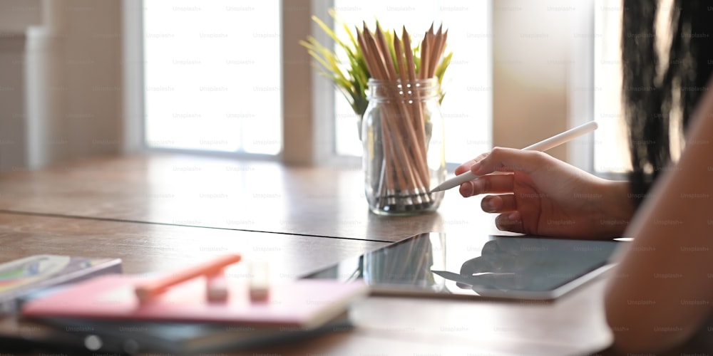 Side shot of creative woman working with computer tablet and stylus pen while sitting at the wooden working desk that surrounded by stationary over sitting room windows as background.