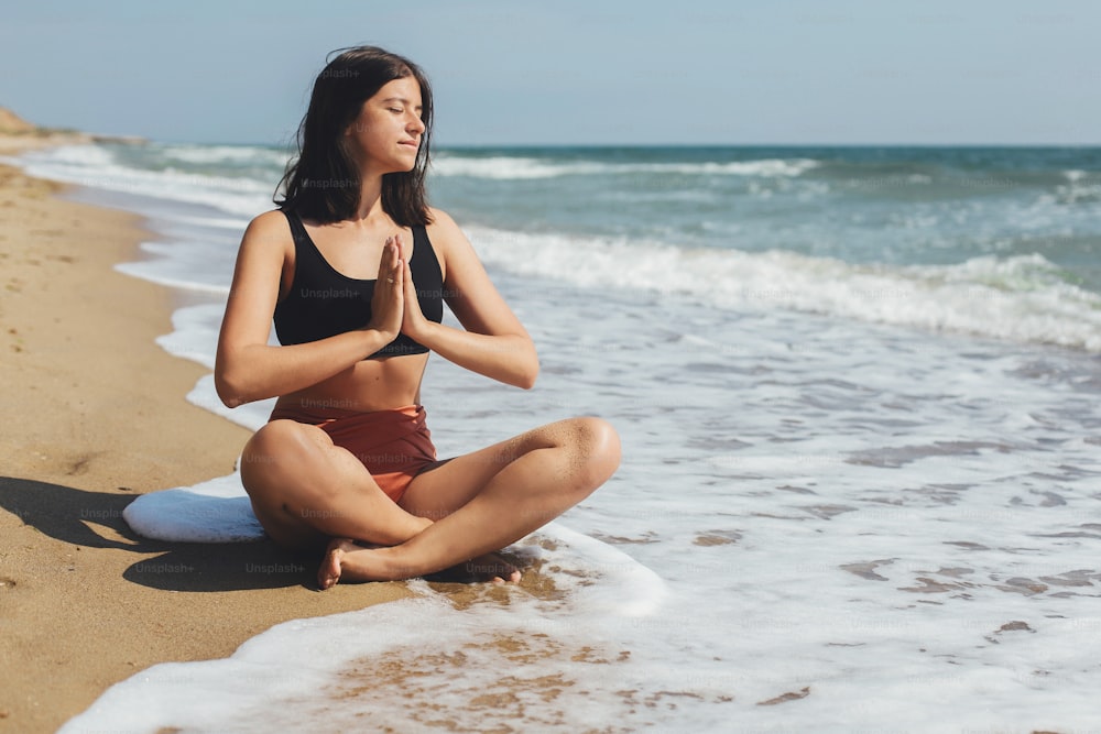 Jeune belle femme pratiquant le yoga sur la plage, assise sur le sable et méditant. Concept de santé mentale et d’autosoins. Fille heureuse se relaxant au bord de la mer pendant les vacances d’été