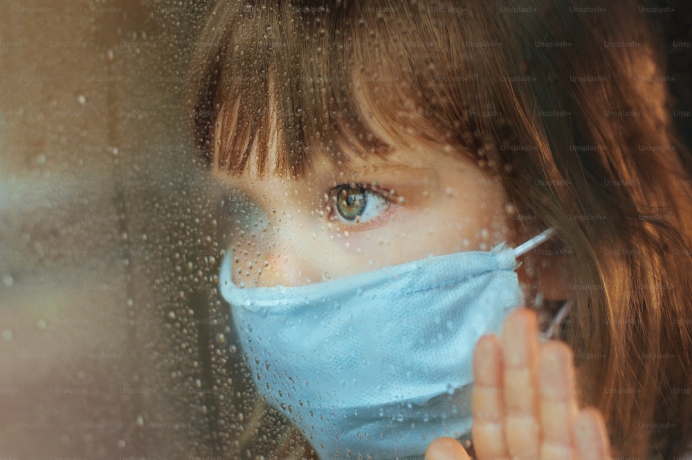 Little girl in face mask looking in the window glass with rain drops. Selective focus on the eyes. Social isolation stay at home during Pandemic COVID-19 concept.