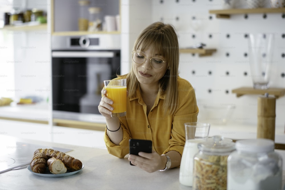 Belle femme buvant du jus d’orange dans la cuisine. Jeune femme lisant les nouvelles en ligne.