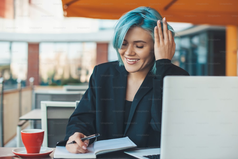 파란 머리를 가진 열심히 일하는 백인 여성은 식당에 앉아 컴퓨터 근처에서 차를 마시는 동안 무언가를 쓰고 있다