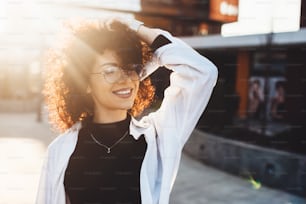 Caucasian charming woman with curly hair touching her head while posing with eyeglass outside