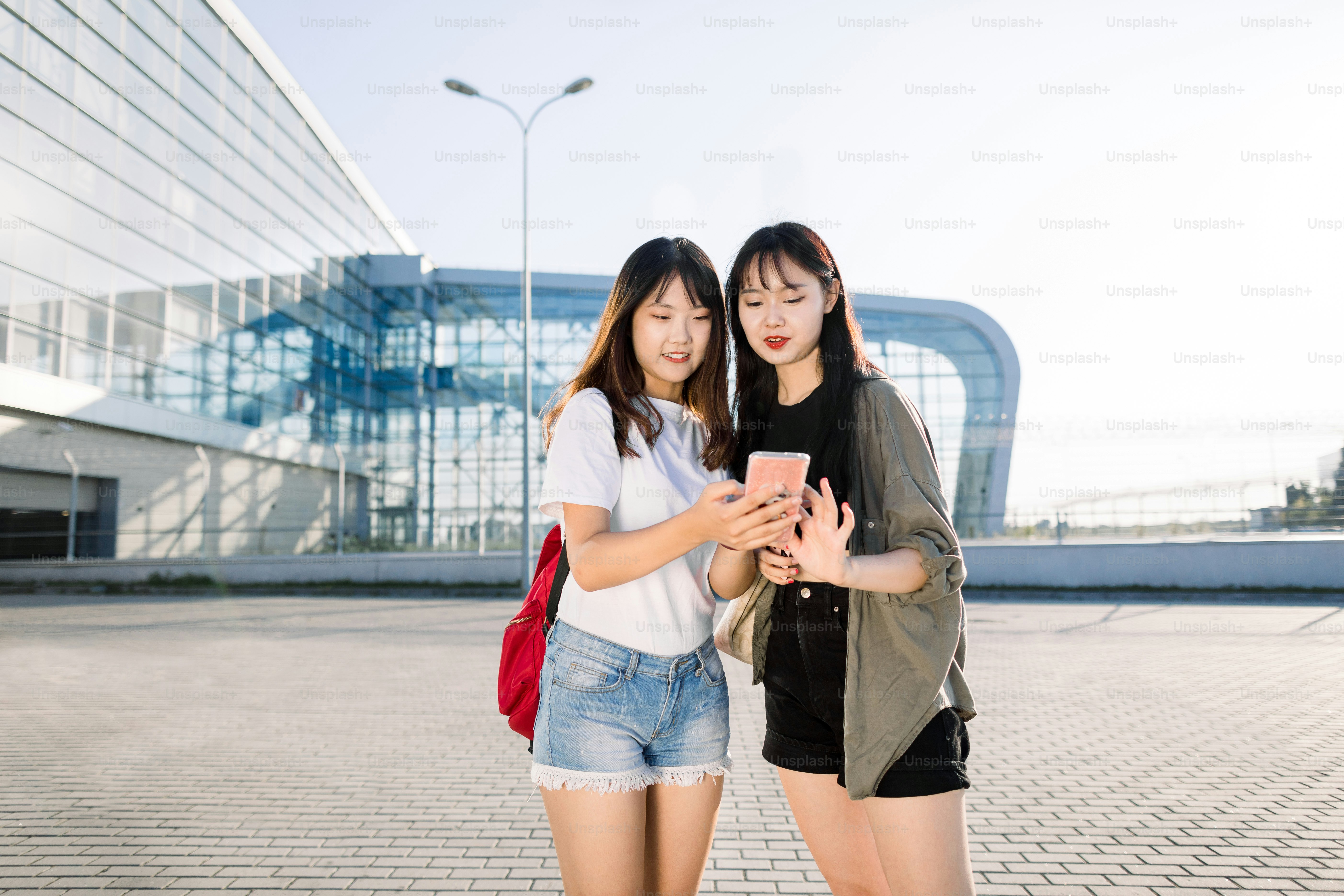Dos chicas asiáticas jóvenes y bonitas de pie frente al edificio del aeropuerto y escribiendo un mensaje o haciendo el check-in en el teléfono inteligente antes de su vuelo conjunto