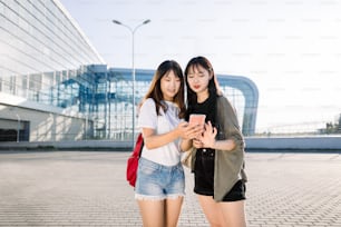 Two pretty young asian girls standing in front of airport building and typing message or making check-in on smartphone before their joint flight