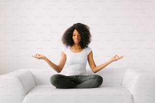 Eine afroamerikanische junge Frau sitzt im Lotussitz auf einem weißen Bett.