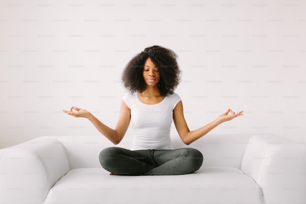 An African American young women sitting in the lotus position on white bed.