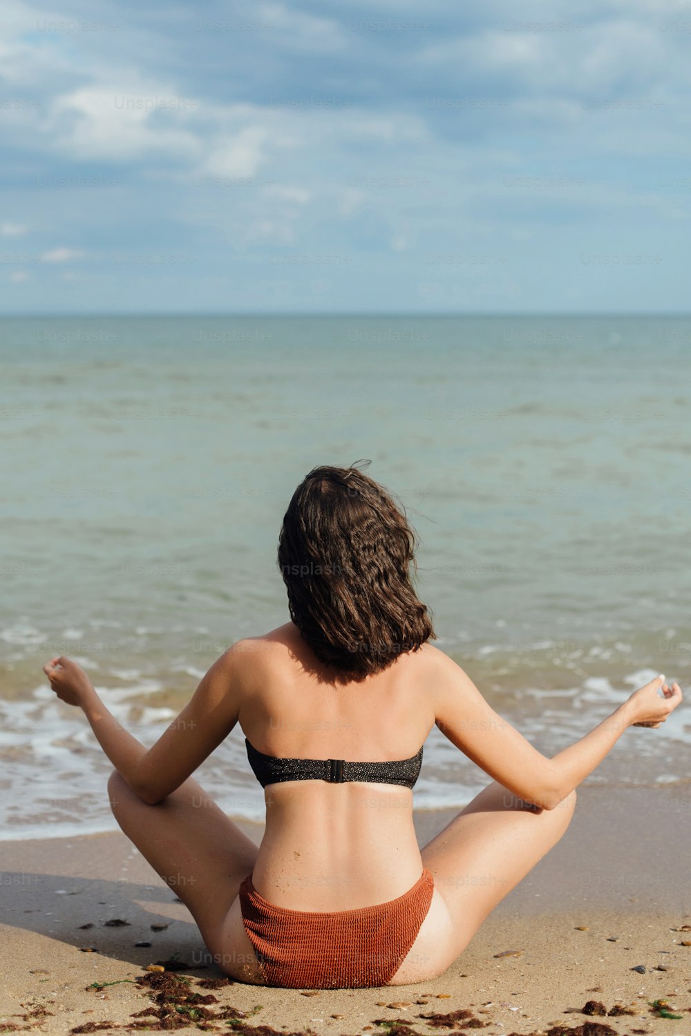 Jeune femme en forme pratiquant le yoga sur la plage, assise sur le sable et regardant les vagues de la mer. Fille méditant pendant les vacances d’été. Concept de santé mentale et d’autosoins
