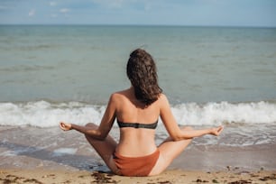 Mental health and self care concept. Young fit woman practicing yoga on the beach, sitting on sand and looking at sea waves. Girl meditating on summer vacation.