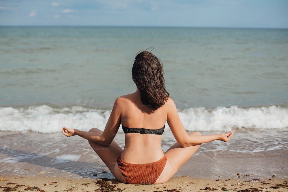 Concetto di salute mentale e cura di sé. Giovane donna in forma che pratica yoga sulla spiaggia, seduta sulla sabbia e guardando le onde del mare. Ragazza che medita sulle vacanze estive.