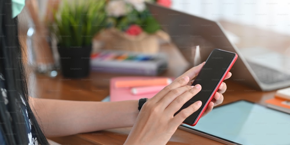 Cropped image of beautiful woman hands holding and using a blank screen smartphone while sitting at the working desk over comfortable working room as background.