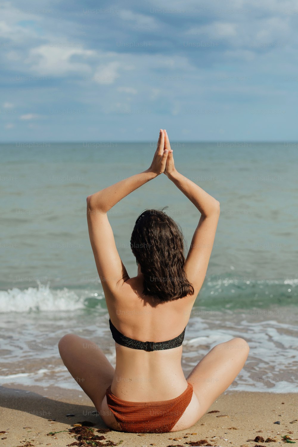 Young fit woman practicing yoga on the beach, sitting on sand and looking at sea waves. Girl meditating on summer vacation. Mental health and self care concept