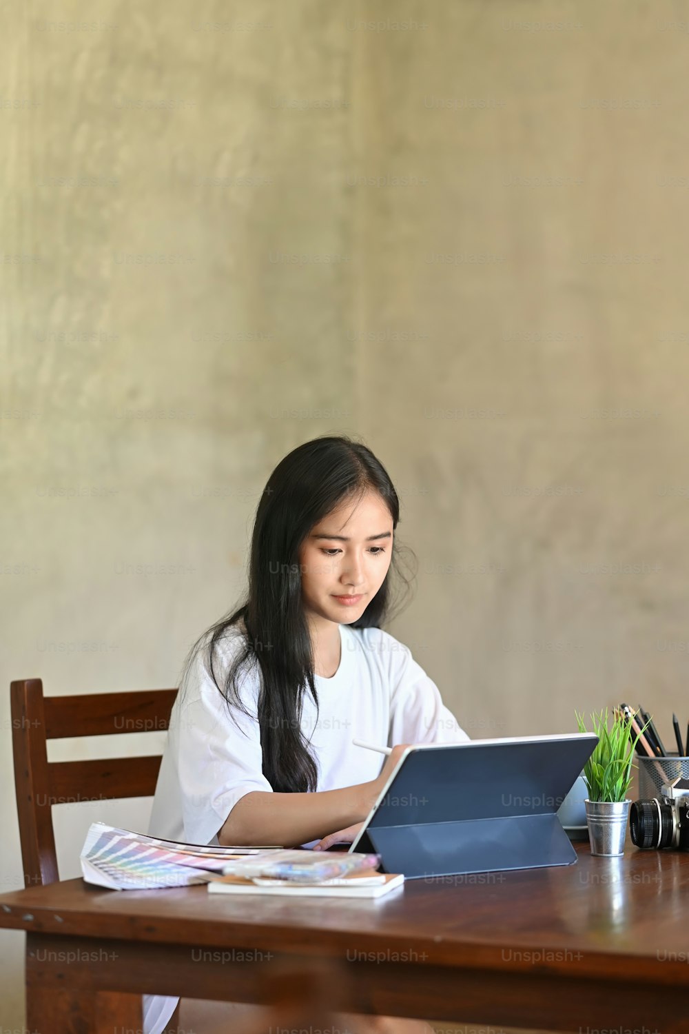 Foto einer Kostümdesignerin, die mit einem Eingabestift auf einem Computertablett skizziert, während sie am hölzernen Schreibtisch über einer bequemen Bürozementwand als Hintergrund sitzt.
