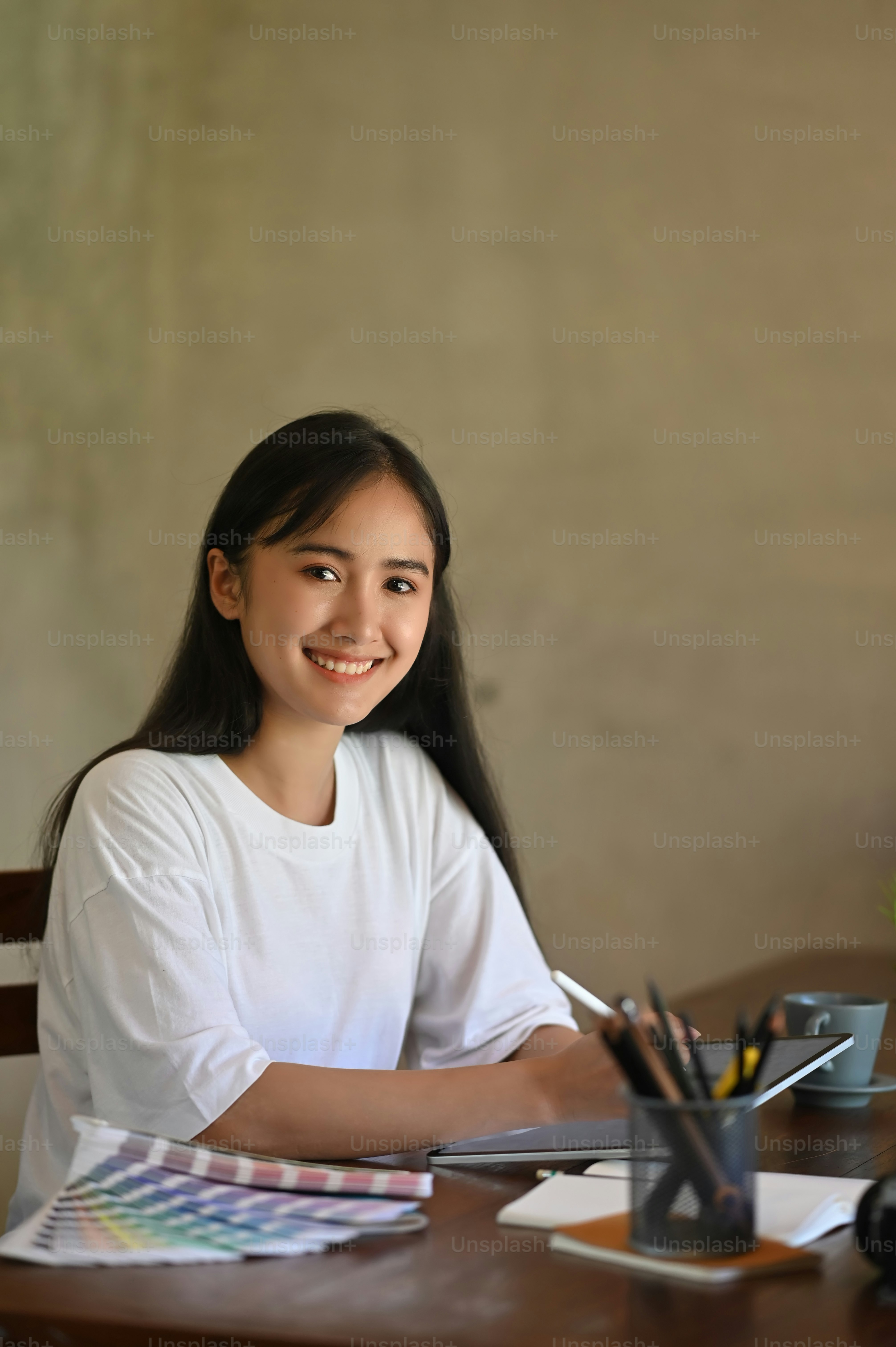 Photo of costume designer woman sketching on computer tablet by using a stylus pen while sitting at the wooden working desk over comfortable office cement wall as background.