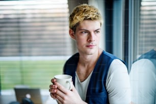 Young man with cup of coffee indoors at home, standing by window and looking out.