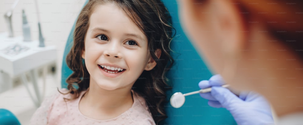 歯医者さんに見て微笑む巻き毛の小さな女の子は、検査後に歯医者さんに微笑みます