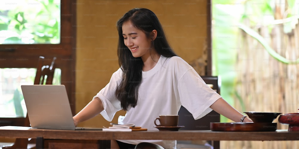 Photo of beautiful woman stretching her body while sitting and relaxing in front her computer laptop that putting on wooden working desk and surrounded by coffee cup and stack of notebook.