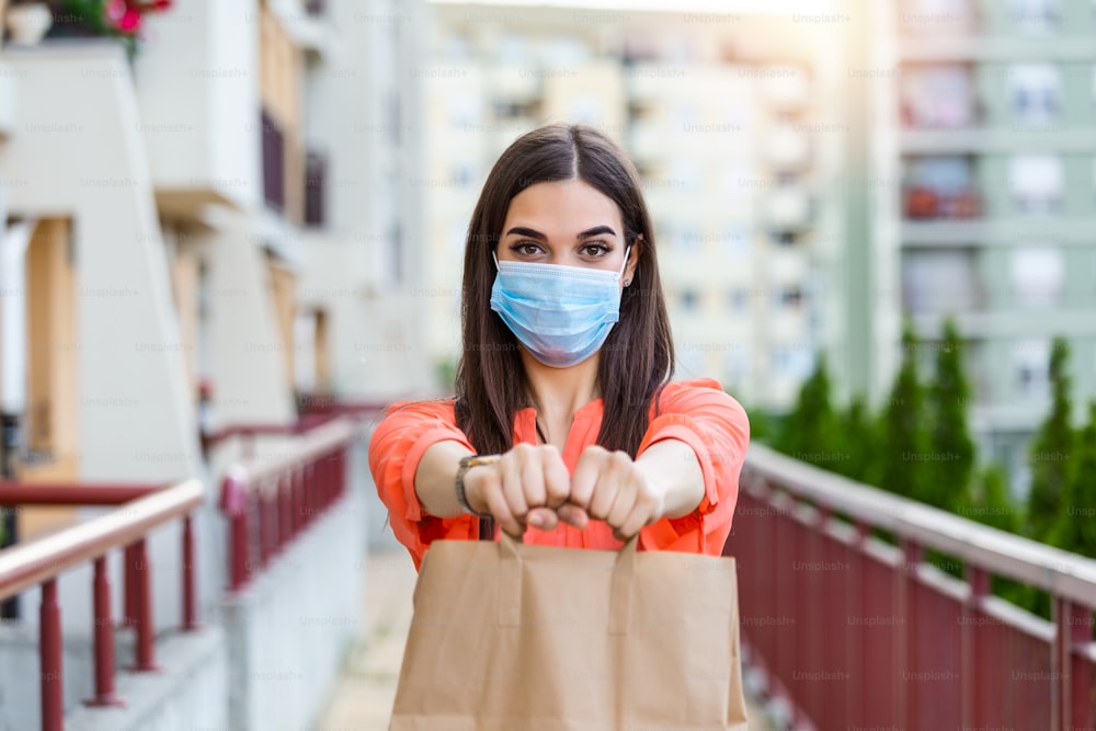 Covid 19、コロナウイルスのパンデミック中の商品の非接触型配送。フードデリバリーの配達員は、大きな紙袋を手に持っています。医療用マスクを着けて食べ物を配達する女性。
