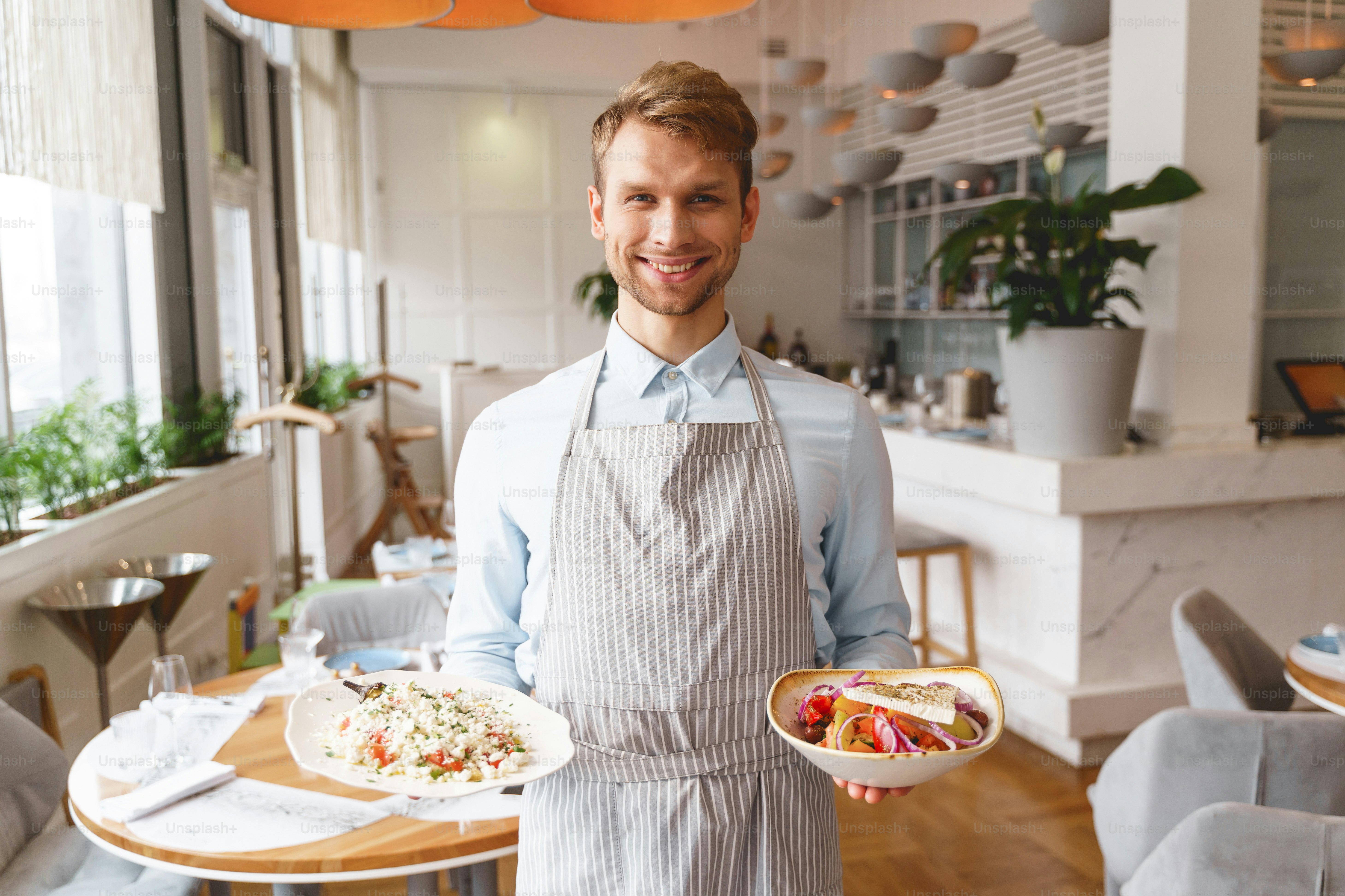 Handsome young man in apron looking at camera and smiling while holding delicious fresh salads